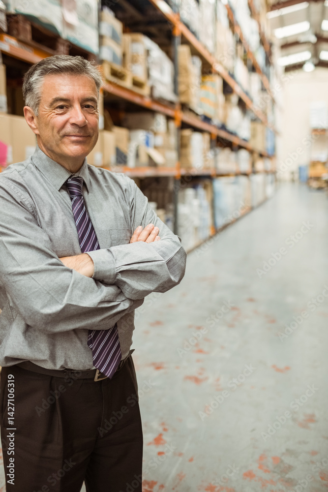 Mature man standing with arms crossed in warehouse aisle, with metal shelves and boxes, copy space
