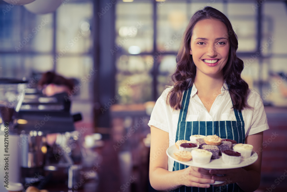 Young female barista holding plate of mixed cupcakes in cafe with espresso machine, copy space