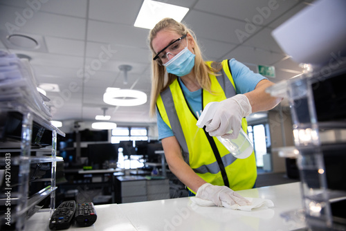 Woman wearing safety vest cleaning office desk with disinfectant bottle and cloth in modern office