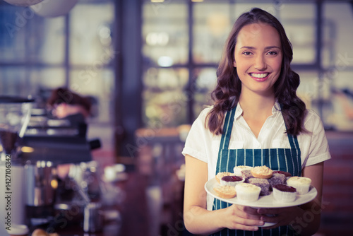 Obraz na płótnie Young female barista holding plate of mixed cupcakes in cafe with espresso machine, copy space w salonie