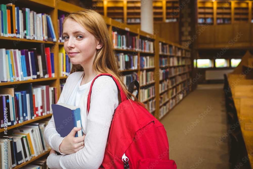Teenage girl standing in library aisle surrounded by bookshelves holding two books and red backpack