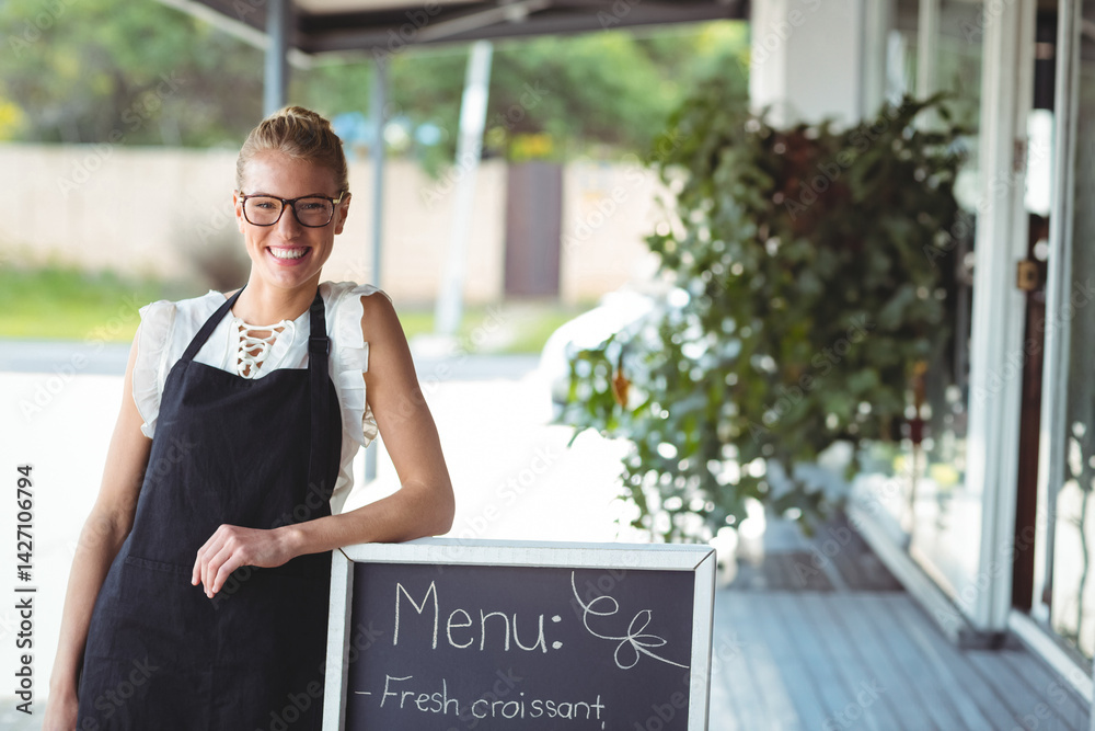 Woman smiling and leaning on chalkboard menu sign under cafe awning, wearing apron and glasses