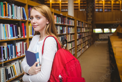 Obraz na płótnie Teenage girl standing in library aisle surrounded by bookshelves holding two books and red backpack w salonie