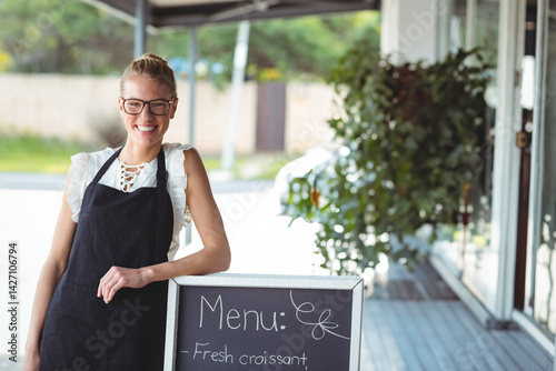 Obraz na płótnie Woman smiling and leaning on chalkboard menu sign under cafe awning, wearing apron and glasses w salonie