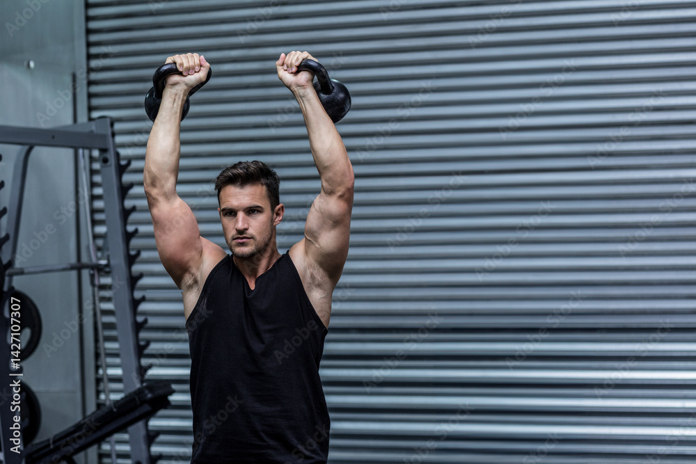 Man lifting kettlebells overhead at industrial-style gym, with weight rack and rubber flooring