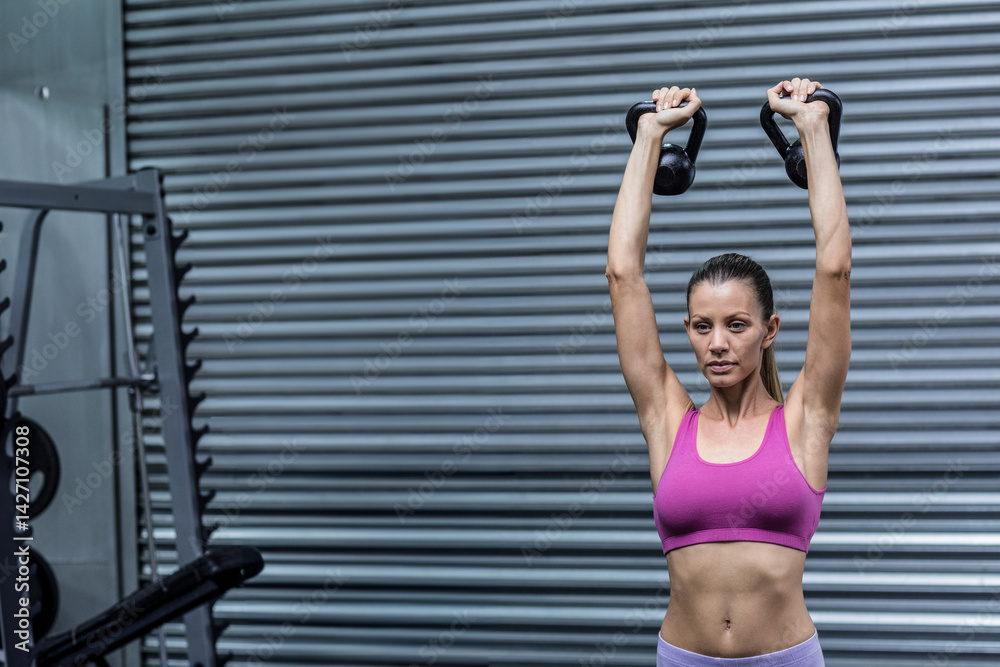 Woman lifting kettlebells overhead in gym with squat rack and loaded weight plates, copy space