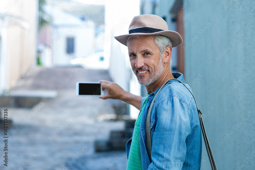 Senior man standing in sunlit stone-paved alley, holding smartphone and wearing fedora, copy space