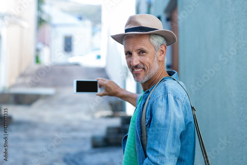 Senior man standing in sunlit stone-paved alley, holding smartphone and wearing fedora, copy space