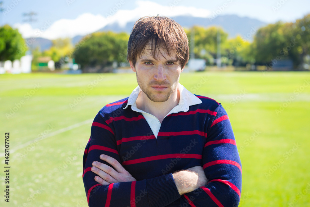 Man standing at center of grassy sports field under sun, crossing arms and gazing at camera