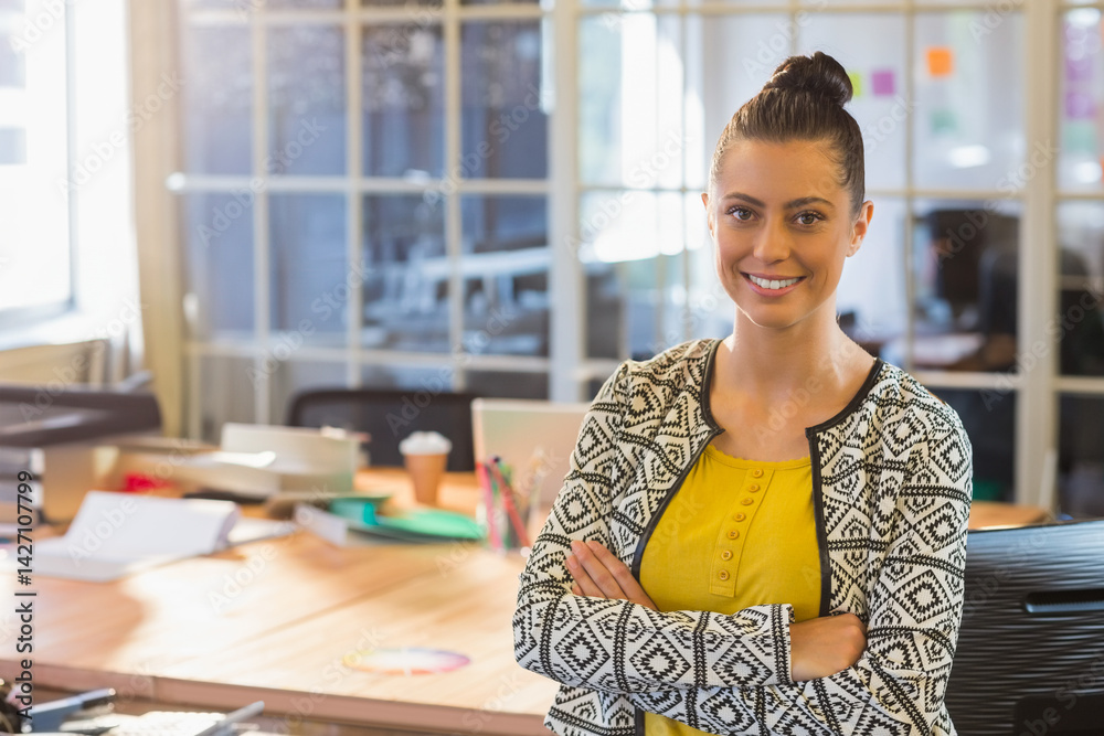 Woman standing with arms crossed in open-plan office, with laptop and coffee cup, copy space