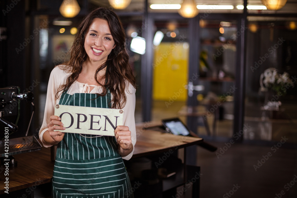 Young woman wearing green apron holding OPEN sign and smiling in café beside espresso machine