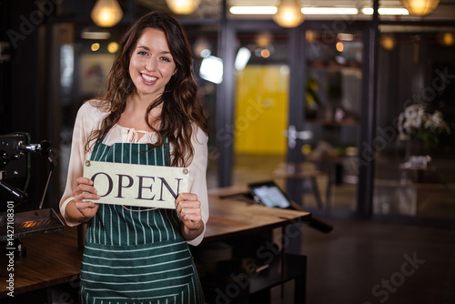 Obraz na płótnie Young woman wearing green apron holding OPEN sign and smiling in café beside espresso machine w salonie