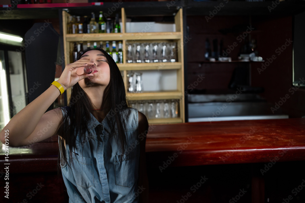 Asian woman drinking from shot glass at bar counter in dimly lit bar, with bar shelves