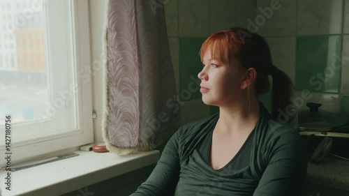A young woman with red hair sits quietly by a window, lost in thought while observing the world outside