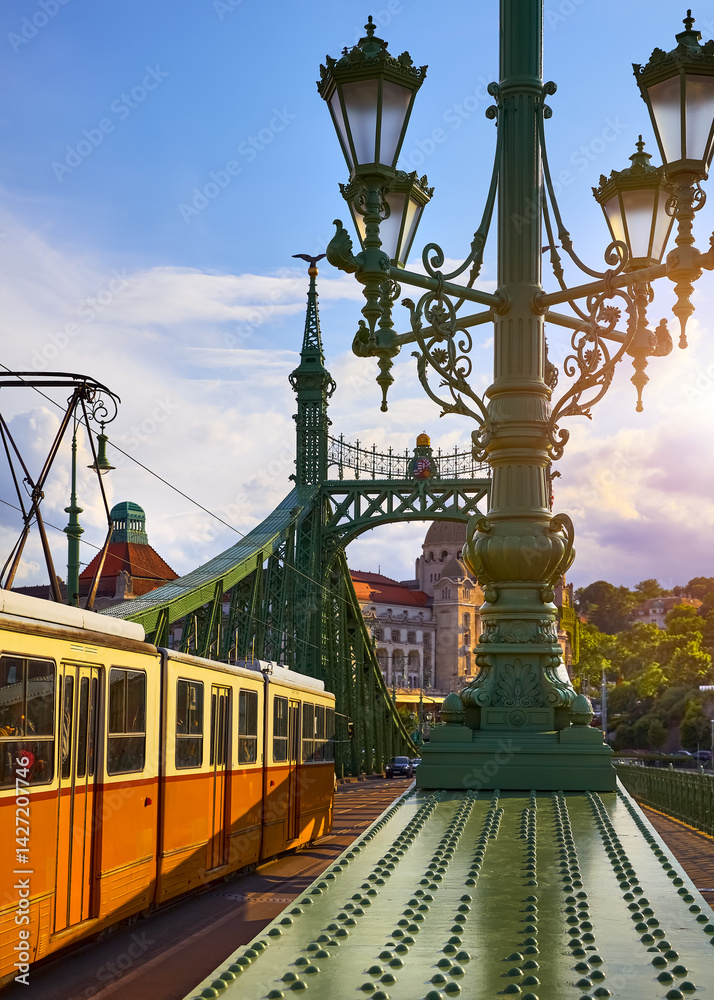 Budapest, Hungary. Old tram on Freedom bridge with sunlight in sunset sky, clouds and pole with street lamps, evening urban landscape. Travel Europe.