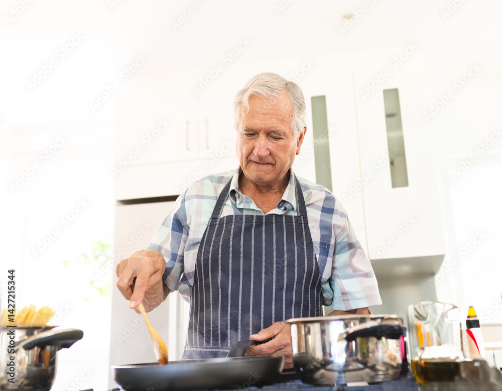 Senior man stirring food in frying pan in bright modern kitchen, with pot and wooden spatula Senior man stirring food in frying pan in bright modern kitchen, with pot and wooden spatula