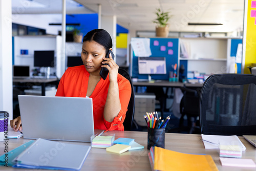 Mid adult woman calling on smartphone and focusing on laptop at modern office with coffee cup