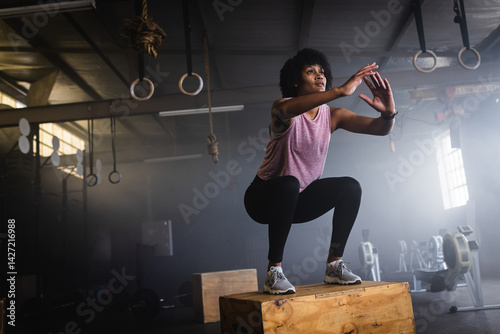 young African American woman lowering into squat on plyo box in industrial gym, copy space photo