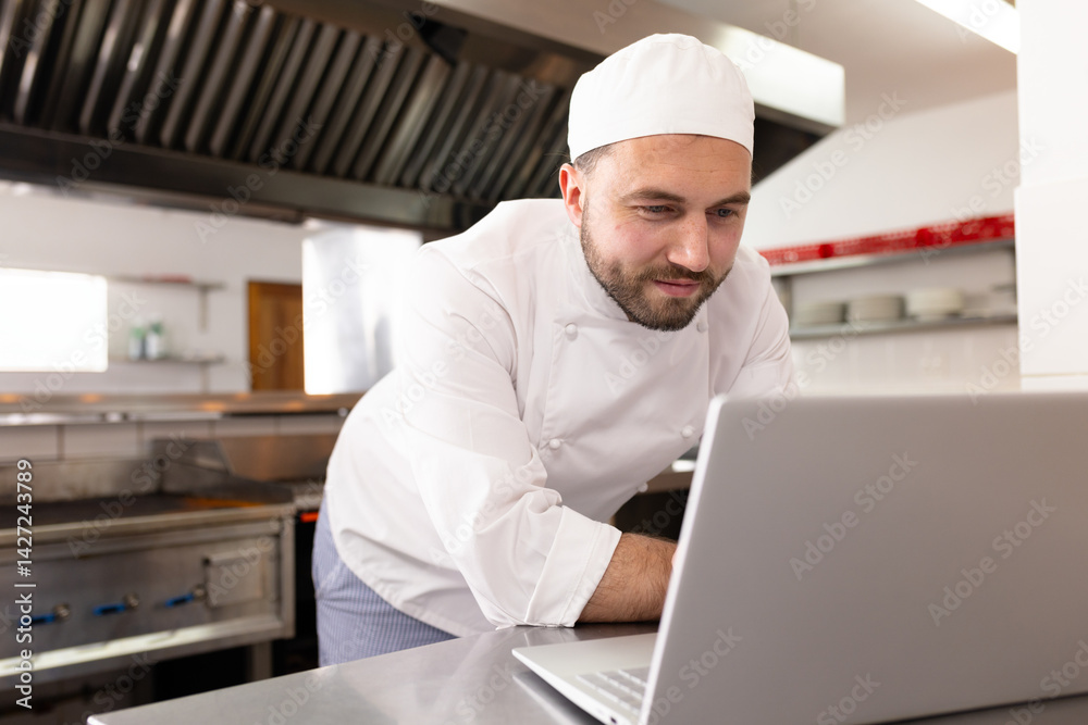 Male chef leaning over steel counter checking silver laptop in commercial kitchen, copy space Male chef leaning over steel counter checking silver laptop in commercial kitchen, copy space