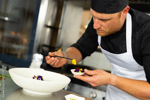 Male chef placing yellow edible flowers into serving bowl in busy kitchen, using black tweezers