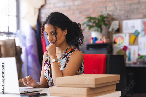 Woman working on laptop in creative studio while sorting mailer boxes smartphone sketches swatches