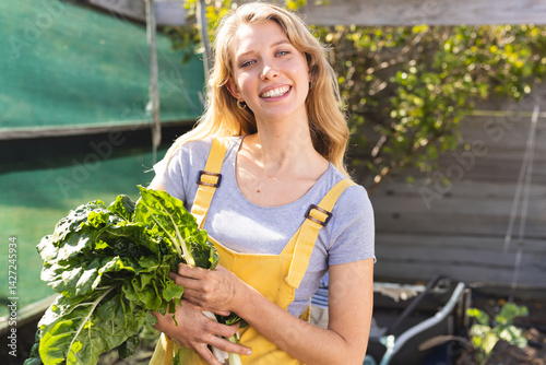 Woman wearing yellow overalls and holding bundle of leafy greens in garden, with green shade cloth