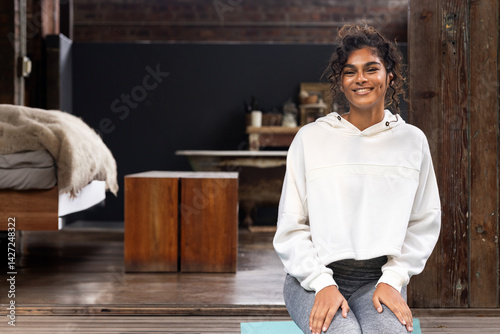Multiracial woman sitting on teal yoga mat in loft style room, smiling at camera, copy space