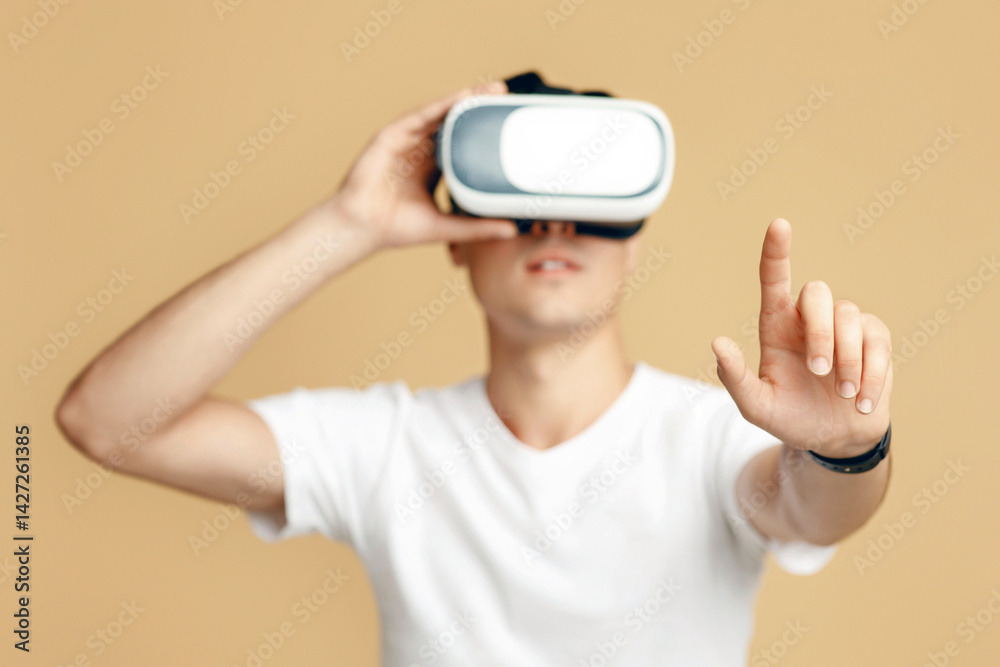 Man in vr glasses, watching 360 degree video with virtual reality headset. Concentrated young man student in white t-shirt touches blank space with finger isolated on sand background, studio shot Man in vr glasses, watching 360 degree video with virtual reality headset. Concentrated young man student in white t-shirt touches blank space with finger isolated on sand background, studio shot