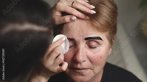 A cosmetologist paints the eyebrows of an elderly woman.