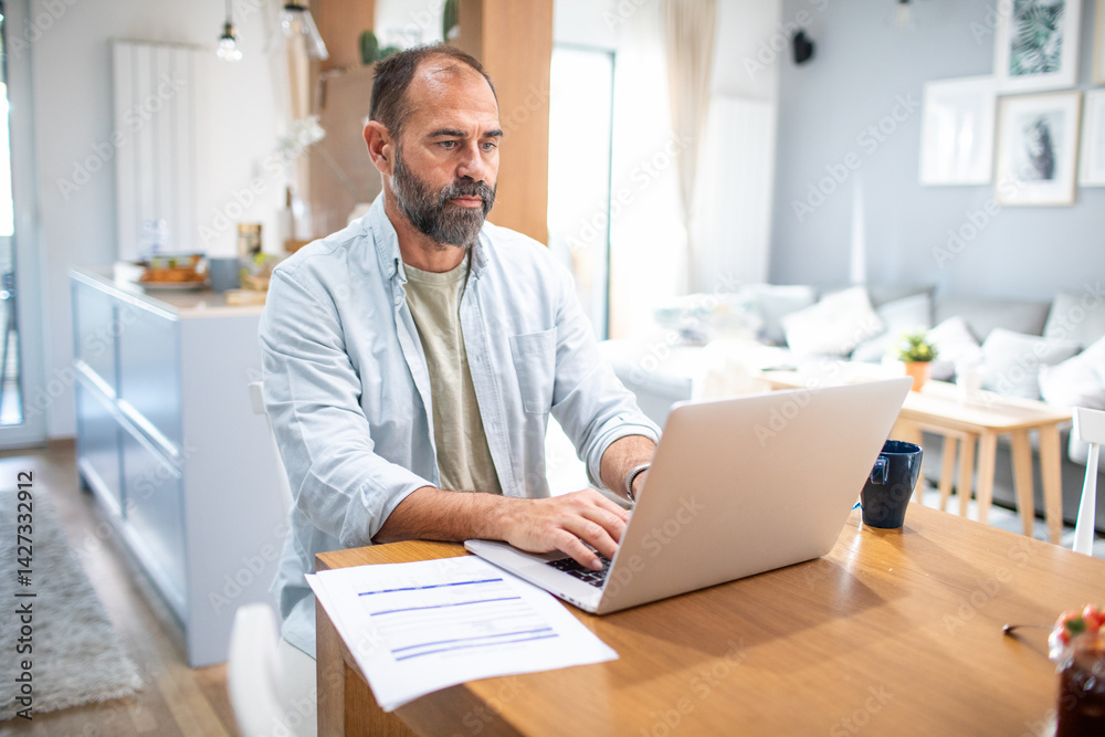 Middle aged man working from home on laptop at kitchen table Middle aged man working from home on laptop at kitchen table