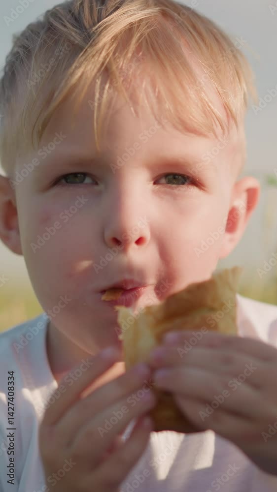 A close-up shot of a young boy wearing a white top, sitting on a grass field while eating. His expression reflects enjoyment as he savors his food in the open air A close-up shot of a young boy wearing a white top, sitting on a grass field while eating. His expression reflects enjoyment as he savors his food in the open air
