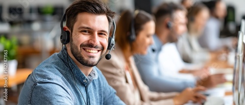 A smiling man in a headset works in a busy office environment surrounded by colleagues at computers.