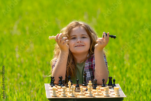 Kid plays chess. A clever blonde child thinks and playing chess. Kid focuses on chessboard. The child make checkmate. A smart kid plans chess. Child sits on grass enjoys chess. The board logic game.