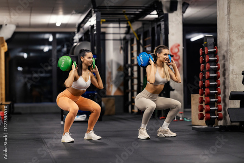 Two strong fit sportswomen with power bags doing squats at gym.