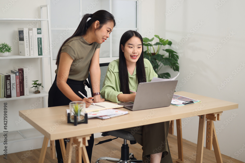 Portrait of Asian woman is standing writing in a notebook while discussing ideas with a colleague sitting in front of a laptop. Portrait of Asian woman is standing writing in a notebook while discussing ideas with a colleague sitting in front of a laptop.