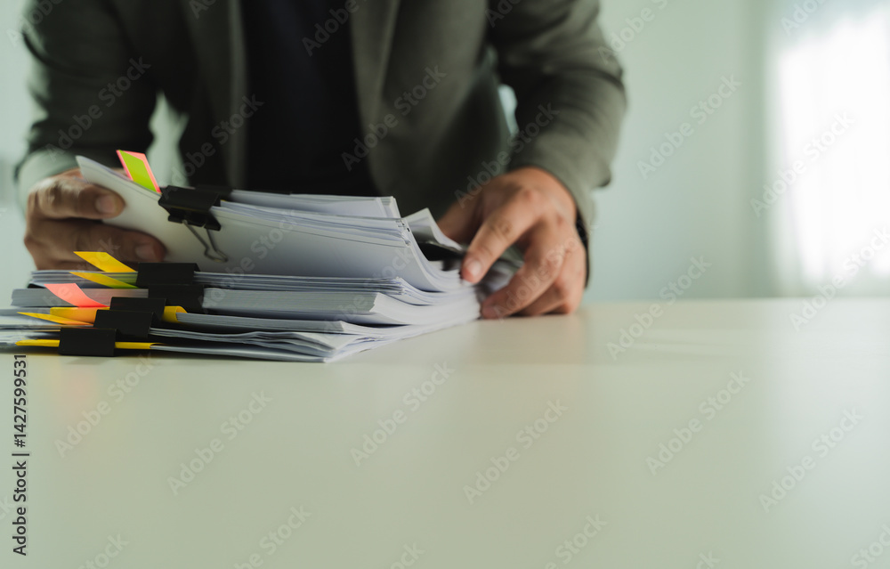 Stacks of paper files and checking document businessman checks financial documents on his laptop, managing corporate reports and legal paperwork at his office desk.