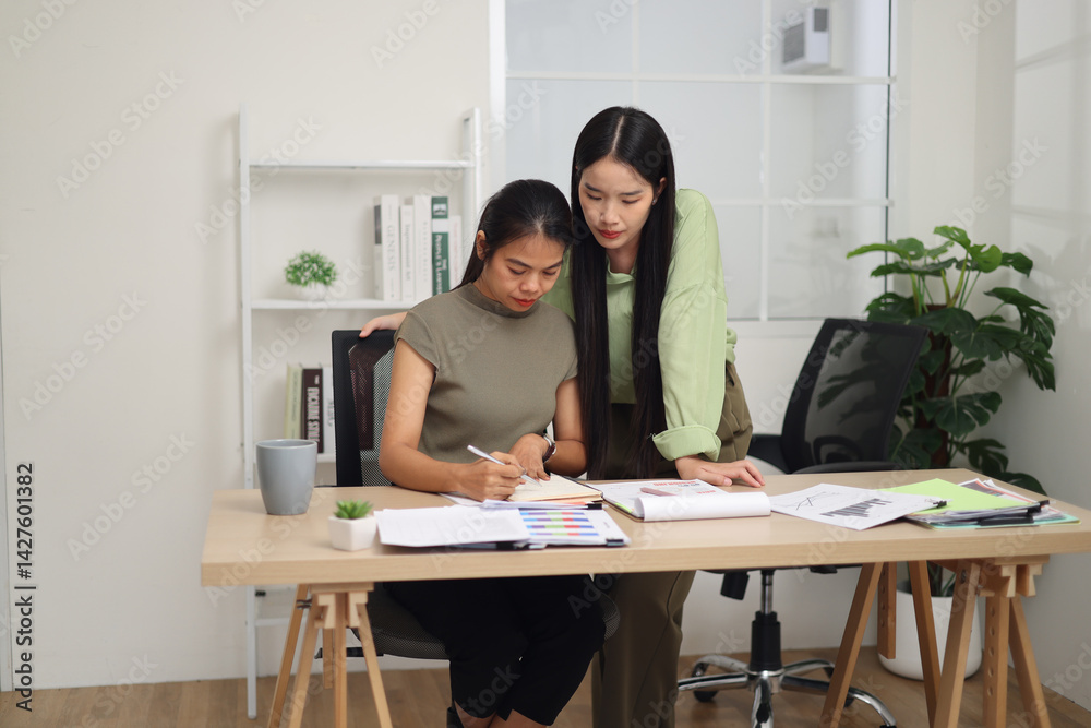 Portrait of Two female colleagues are working together in an office. One stands explaining the work, while the other sits and listens with interest.
