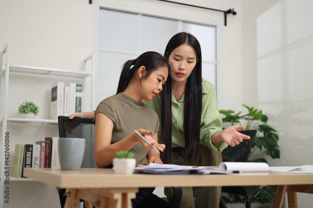 Portrait of Two female colleagues are working together in an office. One stands explaining the work, while the other sits and listens with interest.