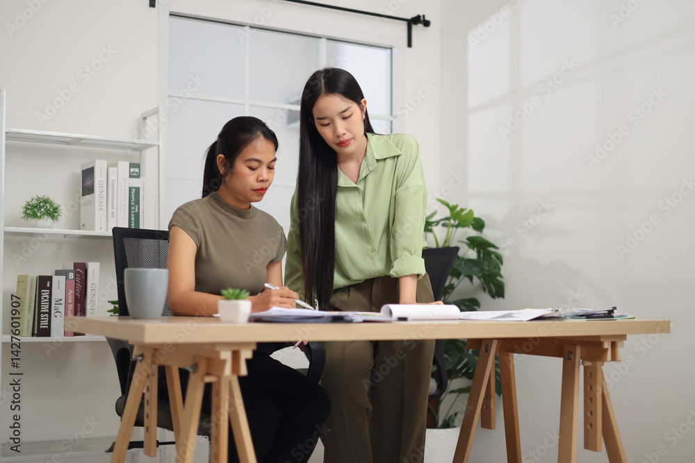 Low angle view shot, Two female colleagues are working together in an office. A young woman is standing, talking and exchanging ideas with another colleague who is sitting.
