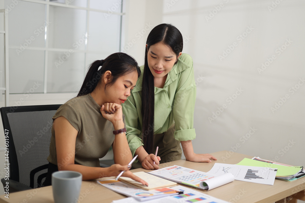 Portrait of Two female colleagues are working together in an office. A young woman is standing, talking and exchanging ideas with another colleague who is sitting.