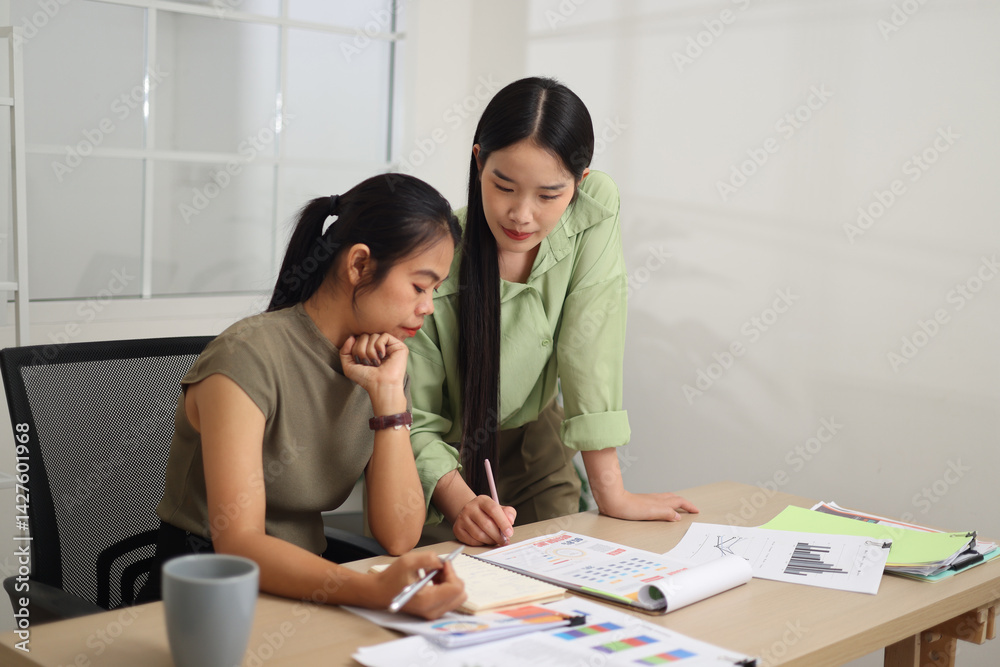 Portrait of Two female colleagues are working together in an office. One stands explaining the work, while the other sits and listens with interest.