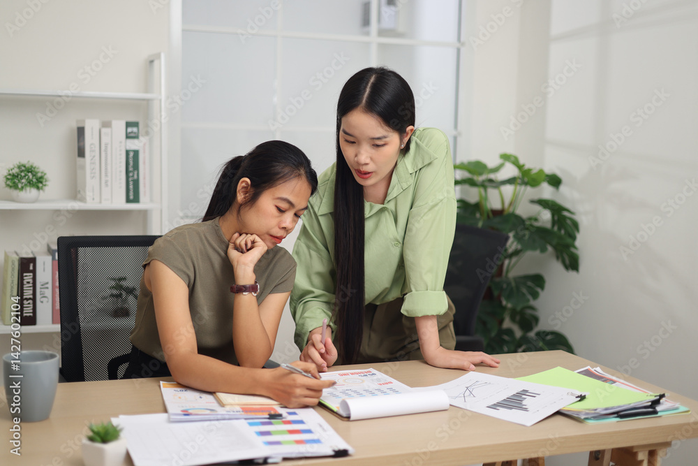 Portrait of Two female colleagues are working together in an office. One stands explaining the work, while the other sits and listens with interest.