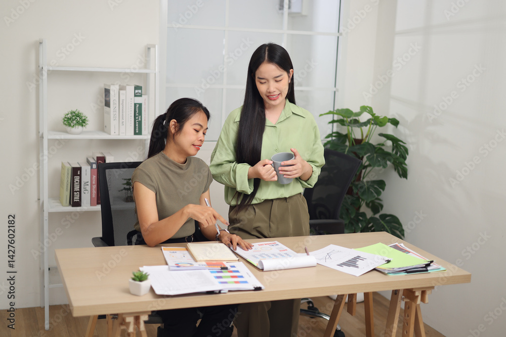 High angle view shot, Two female colleagues are working together in an office. A young woman is standing holding a cup of coffee and exchanging ideas with another colleague who is sitting.