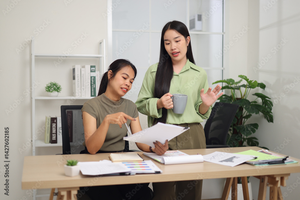 Portrait of Two female colleagues are working together in an office. A young woman is standing holding a cup of coffee and exchanging ideas with another colleague who is sitting.