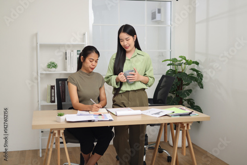 Obraz na płótnie Portrait of Two female colleagues are working together in an office. A young woman is standing holding a cup of coffee and exchanging ideas with another colleague who is sitting. w salonie