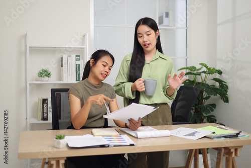 Obraz na płótnie Portrait of Two female colleagues are working together in an office. A young woman is standing holding a cup of coffee and exchanging ideas with another colleague who is sitting. w salonie