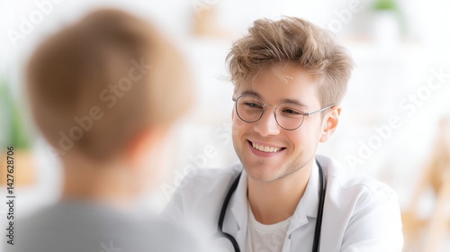 A caring young doctor with glasses is smiling and talking to a child patient in a clinic.
