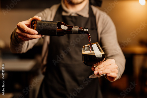 Photos A bartender in an apron pours dark beer from a brown bottle into a tulip glass under warm indoor lighting