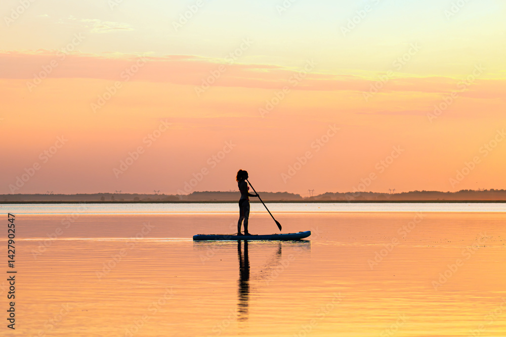 woman stands on a sup board with a paddle on the rowing channel against the background of a yellow sunset. Active water sports. A sexy woman is engaged in rowing.