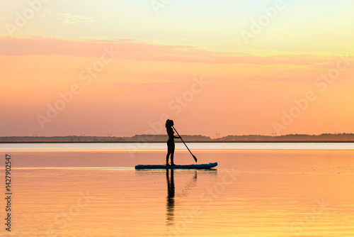 Obraz na płótnie woman stands on a sup board with a paddle on the rowing channel against the background of a yellow sunset. Active water sports. A sexy woman is engaged in rowing. w salonie